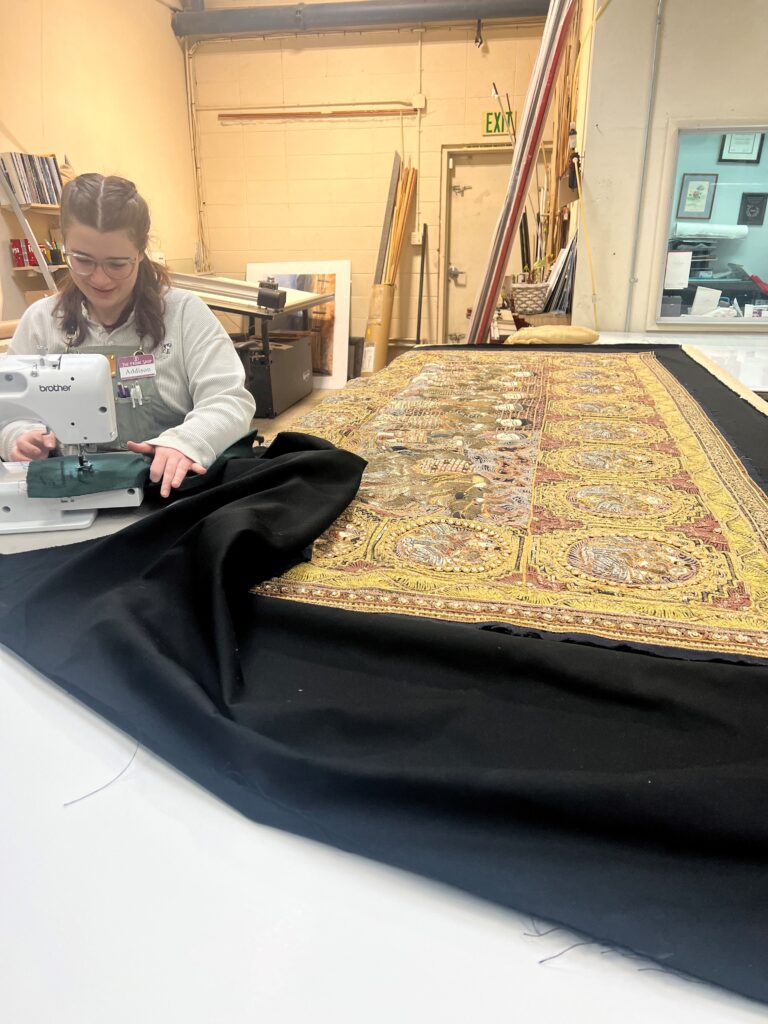 Tapestry A woman sewing on a machine next to a large piece of fabric.